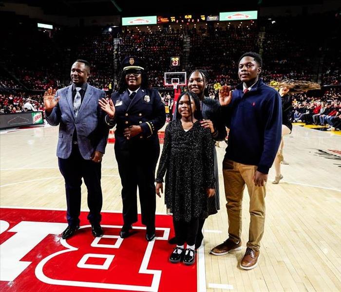 A group of five people, including a uniformed officer, stand together on a basketball court with a crowd in the background. 